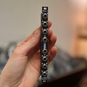 May include: A black electronic module with multiple input and output jacks. The module has the text "MULT" at the top and "INPUT" in the middle. The bottom of the module has the text "TEMBRA KAXIMIA".