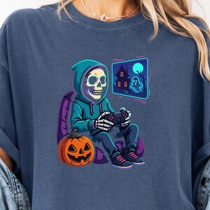 May include: Navy blue t-shirt featuring a Halloween-themed graphic. The design shows a skeleton wearing a hoodie and playing a video game, with a jack-o'-lantern and a haunted house on a screen. The shirt is a comfortable, casual style.