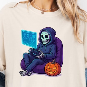 May include: Cream-colored t-shirt featuring a cartoon skeleton in a blue hoodie playing a video game. The skeleton sits on a purple beanbag chair next to an orange jack-o'-lantern. The video game screen is blue. A Halloween-themed graphic tee.
