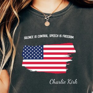 May include: Dark gray t-shirt featuring an American flag design with the text "SILENCE IS CONTROL SPEECH IS FREEDOM" and "Charlie Kirk". The flag is red, white, and blue. The shirt is a casual wear item.