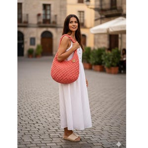 May include: A coral-colored, crocheted hobo bag with a wooden button closure. The bag is carried over the shoulder. The woman is wearing a white dress and beige sandals. The background is a cobblestone street and buildings.