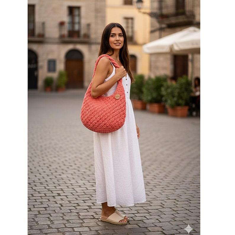 May include: A coral-colored, crocheted hobo bag with a wooden button closure. The bag is carried over the shoulder. The woman is wearing a white dress and beige sandals. The background is a cobblestone street and buildings.