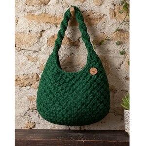 May include: A dark green, handmade crochet bag with a braided handle, hanging against a textured stone wall. The bag has a rounded shape and a small, round wooden tag attached. The bag is approximately 14 inches wide and 12 inches tall.