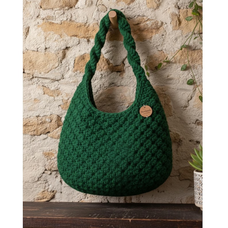 May include: A dark green, handmade crochet bag with a braided handle, hanging against a textured stone wall. The bag has a rounded shape and a small, round wooden tag attached. The bag is approximately 14 inches wide and 12 inches tall.