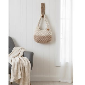 May include: A two-tone, cream and tan crochet handbag hangs from a wooden wall mount. The bag has a rounded shape with a braided handle and a small, round, brown leather tag. A gray armchair and a cream-colored blanket are in the background.