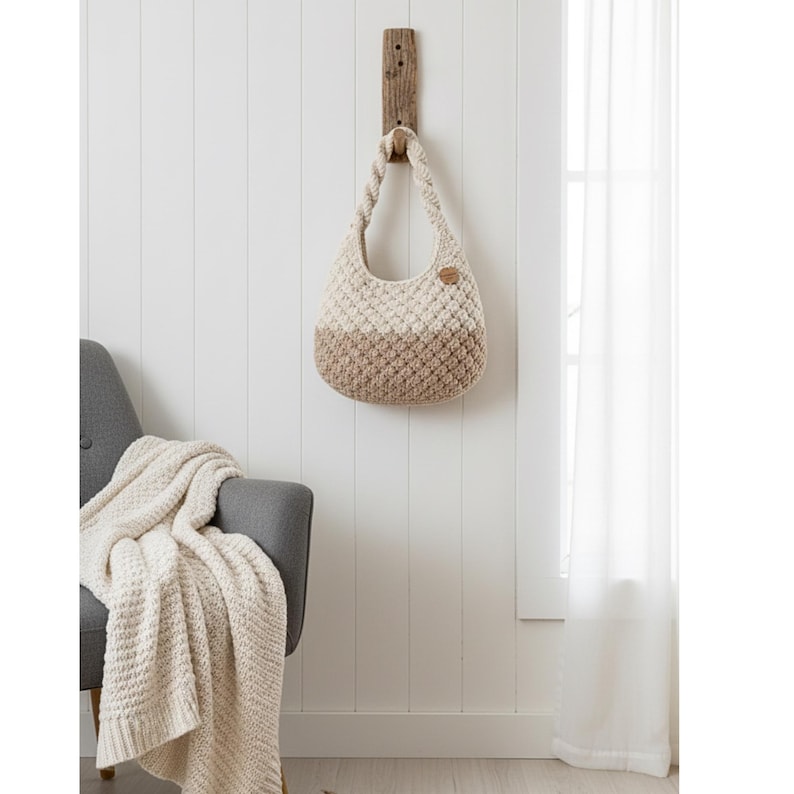 May include: A two-tone, cream and tan crochet handbag hangs from a wooden wall mount. The bag has a rounded shape with a braided handle and a small, round, brown leather tag. A gray armchair and a cream-colored blanket are in the background.