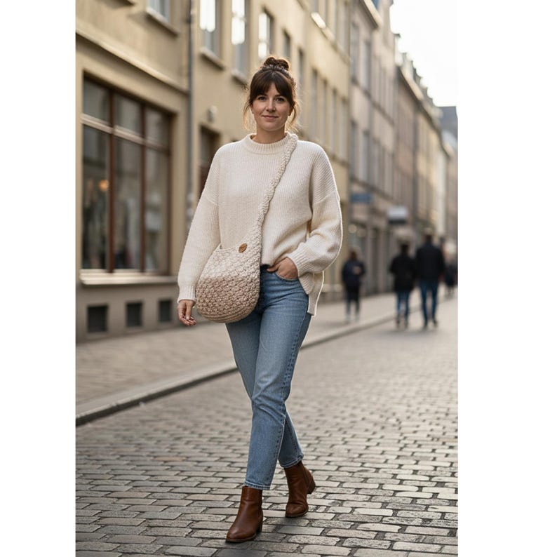 May include: A woman wearing a cream-colored sweater, light blue jeans, and brown leather boots walks down a cobblestone street. She carries a beige and tan crocheted shoulder bag. The bag has a round shape and a long strap.