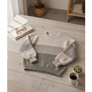 Peut inclure: Un pull rayé crème, gris et vert avec des manches longues. Le pull est fait d'une matière tricotée épaisse. Une petite tasse à café blanche et une petite plante succulente sont à côté du pull sur une surface en bois.