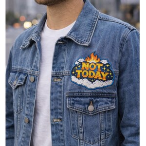 Pode incluir: Casaco de ganga azul claro com uma t-shirt branca. O casaco tem um patch no peito com as palavras "NOT TODAY" em amarelo e laranja, rodeado por nuvens e estrelas. Tem dois bolsos no peito e botões prateados.