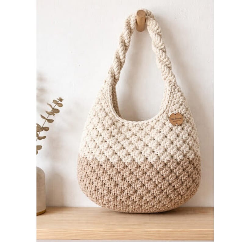 May include: A handmade, cream and tan crochet bag with a braided handle. The bag is hanging on a wooden peg. The bottom half of the bag is tan, and the top half is cream. A small, brown leather tag is attached to the bag.