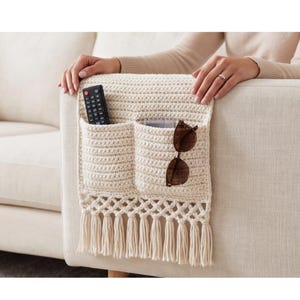 May include: Cream-colored, crocheted couch armrest organizer with two pockets and a fringe. A black remote and brown sunglasses are in the pockets. The organizer is placed on a beige couch armrest.