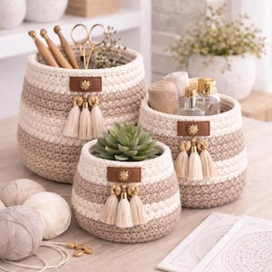 Peut inclure: Ensemble de trois paniers crochetés à la main, de tailles différentes. Les paniers présentent des rayures alternées crème et beige. Chaque panier est orné d'une étiquette en cuir marron, d'un emblème doré et de pompons crème. Un panier contient une plante succulente.