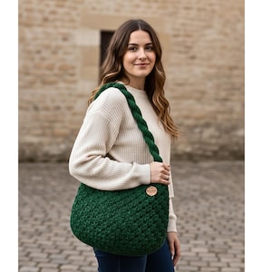 May include: A dark green, crocheted shoulder bag with a braided strap. The bag has a rounded shape and a textured stitch pattern. A small, round wooden tag is attached to the bag. The woman is wearing a cream-colored sweater.