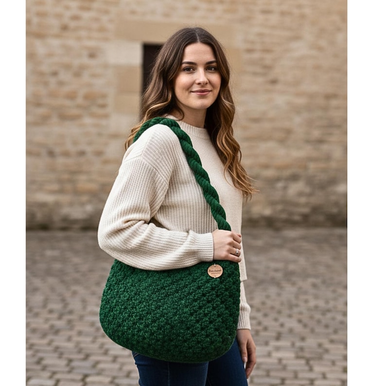 May include: A dark green, crocheted shoulder bag with a braided strap. The bag has a rounded shape and a textured stitch pattern. A small, round wooden tag is attached to the bag. The woman is wearing a cream-colored sweater.