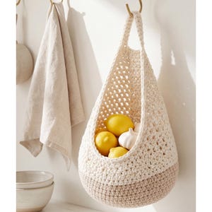 May include: A cream and tan crochet hanging fruit basket filled with yellow lemons and white garlic bulbs. The basket is suspended from a gold hook on a white wall. A beige towel hangs nearby.