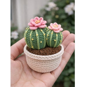 May include: A crocheted cactus in a small pot. The cactus is green with yellow accents and topped with pink flowers. The pot is beige and filled with brown soil. The handmade item is held in a person's hand.