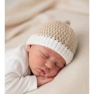 Könnte beinhalten: Ein schlafendes Baby mit einer gehäkelten Mütze in Beige und Weiß. Die Mütze hat ein strukturiertes Muster auf der Krone und einen gerippten weißen Rand. Das Baby trägt einen weißen Strampler und liegt auf einer cremefarbenen Decke.