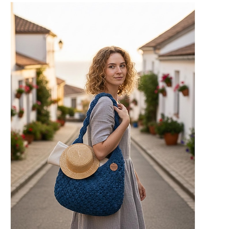 May include: A blue crocheted shoulder bag with a braided strap, holding a straw hat and a white cloth. The bag has a round wooden tag. The woman is wearing a gray dress and standing on a cobblestone street.