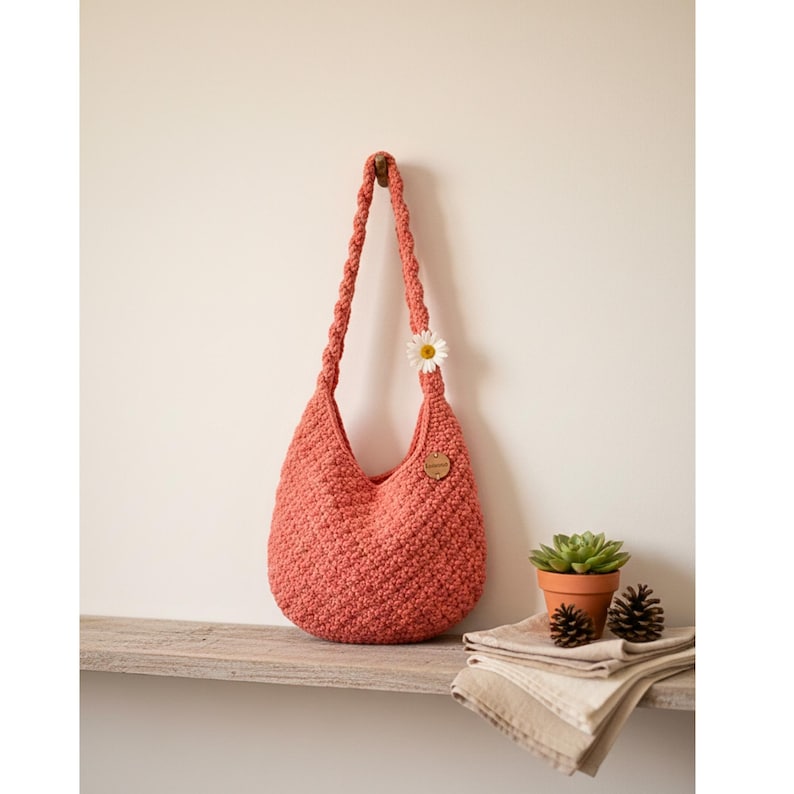 May include: A coral-colored, crocheted hobo bag with a braided strap, adorned with a daisy and a small wooden tag. The bag hangs from a wooden hook on a light-colored wall. A small succulent in a terracotta pot sits on a wooden shelf.