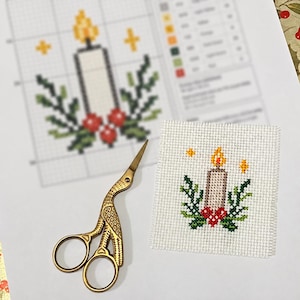 May include: A square piece of white Aida fabric featuring a cross-stitch design of a lit candle with green holly leaves and red berries, accompanied by gold-toned stork-shaped embroidery scissors resting on a printed pattern chart.