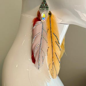 May include: A pair of dangling earrings featuring colorful feathers. The earrings have a decorative top with rhinestones and a hook closure. The feathers are red, light pink, and yellow, with rhinestone accents along the edges.