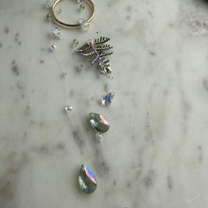 Puede incluir: Un collar delicado e iridiscente con un dije de hoja verde y múltiples cristales en forma de lágrima. El collar está hecho de hilo transparente y tiene un anillo de oro.