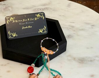 May include: A handcrafted jewelry piece with a gold-colored ring adorned with small purple gems and a decorative copper-colored accent. The piece features beaded strands in green, peach, and red, with two peach-colored beaded orbs. A business card with gold text is also present.