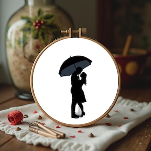 Puede incluir: Un bastidor de bordado de madera con una silueta romántica de una pareja bajo un paraguas. La imagen es en blanco y negro, con la pareja abrazándose. El bastidor está sobre un paño blanco con hilo rojo y herramientas doradas.