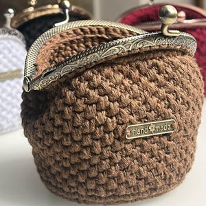 May include: A brown crocheted coin purse with a vintage-style metal clasp and a gold tag that reads "Hand Made".