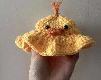 May include: A handmade, yellow crochet duck hat with orange accents. The hat features a round shape with a ruffled brim, black eyes, and an orange beak. A small tuft of orange yarn sits atop the hat. The hat is held in a hand.