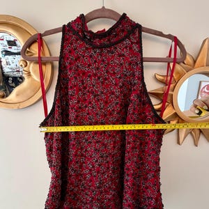 K&ouml;nnte beinhalten: &Auml;rmelloses Oberteil mit hohem Ausschnitt in tiefem Rot, verziert mit einem Muster aus schwarzen und silbernen Perlenbl&auml;ttern. Das Oberteil h&auml;ngt an einem Kleiderb&uuml;gel, mit einem Ma&szlig;band, das seine Breite anzeigt. Das Oberteil hat einen schwarzen Perlenbesatz.