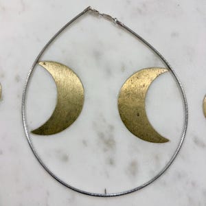 May include: A silver-toned necklace with two gold-colored crescent moon pendants. The necklace is displayed on a white marble surface. The moons have a textured, aged appearance.