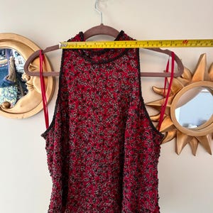 K&ouml;nnte beinhalten: Ein &auml;rmelloses, rotes Oberteil mit schwarzem Perlenbesatz und einem Bl&auml;ttermuster. Das Oberteil h&auml;ngt an einem braunen Kleiderb&uuml;gel. Das Oberteil ist etwa 33 cm breit.