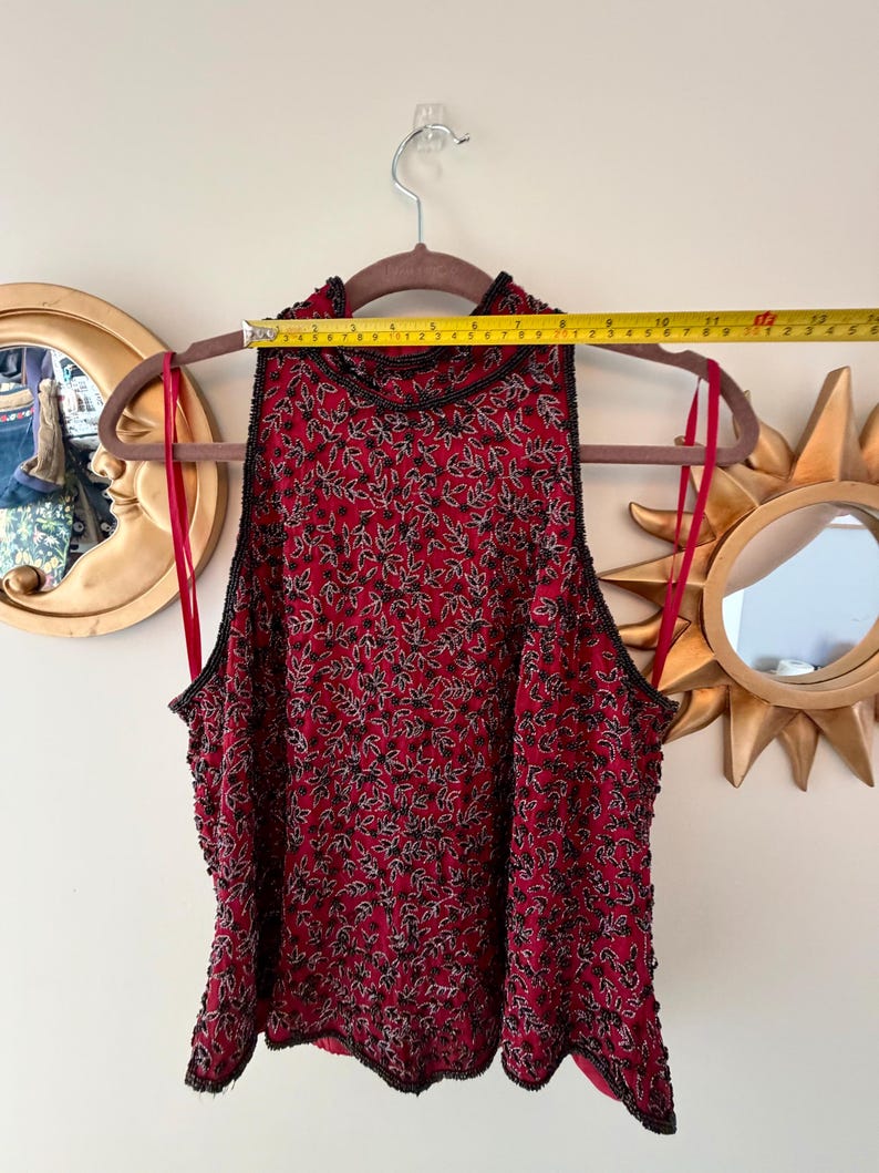 K&ouml;nnte beinhalten: Ein &auml;rmelloses, rotes Oberteil mit schwarzem Perlenbesatz und einem Bl&auml;ttermuster. Das Oberteil h&auml;ngt an einem braunen Kleiderb&uuml;gel. Das Oberteil ist etwa 33 cm breit.