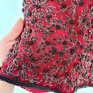 K&ouml;nnte beinhalten: Nahaufnahme eines roten Kleidungsst&uuml;cks mit aufwendiger Perlenarbeit. Das Design zeigt silberne und schwarze Perlen in einem floralen Muster mit Bl&auml;ttern und Beerentrauben. Die R&auml;nder sind mit schwarzen Perlen verziert.