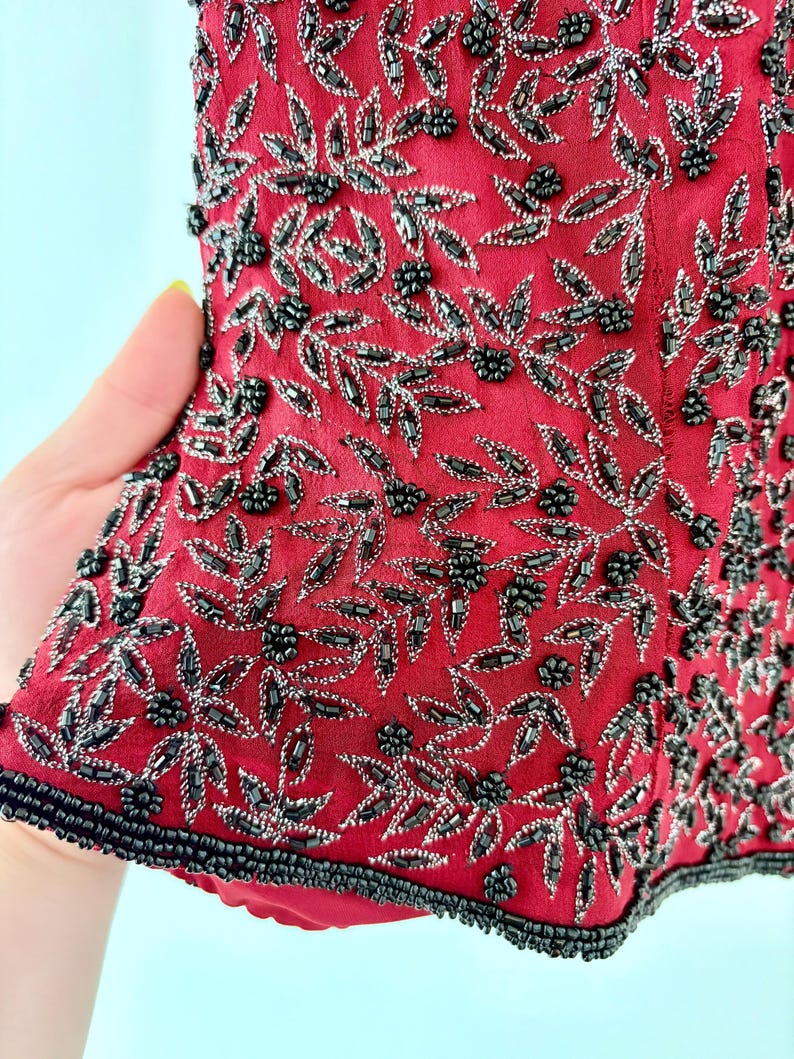 K&ouml;nnte beinhalten: Nahaufnahme eines roten Kleidungsst&uuml;cks mit aufwendiger Perlenarbeit. Das Design zeigt silberne und schwarze Perlen in einem floralen Muster mit Bl&auml;ttern und Beerentrauben. Die R&auml;nder sind mit schwarzen Perlen verziert.
