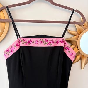 Puede incluir: Vestido negro con tirantes finos y ribete bordado floral rosa. El vestido est&aacute; colgado de una percha marr&oacute;n. El ribete presenta rosas rosas y hojas verdes.
