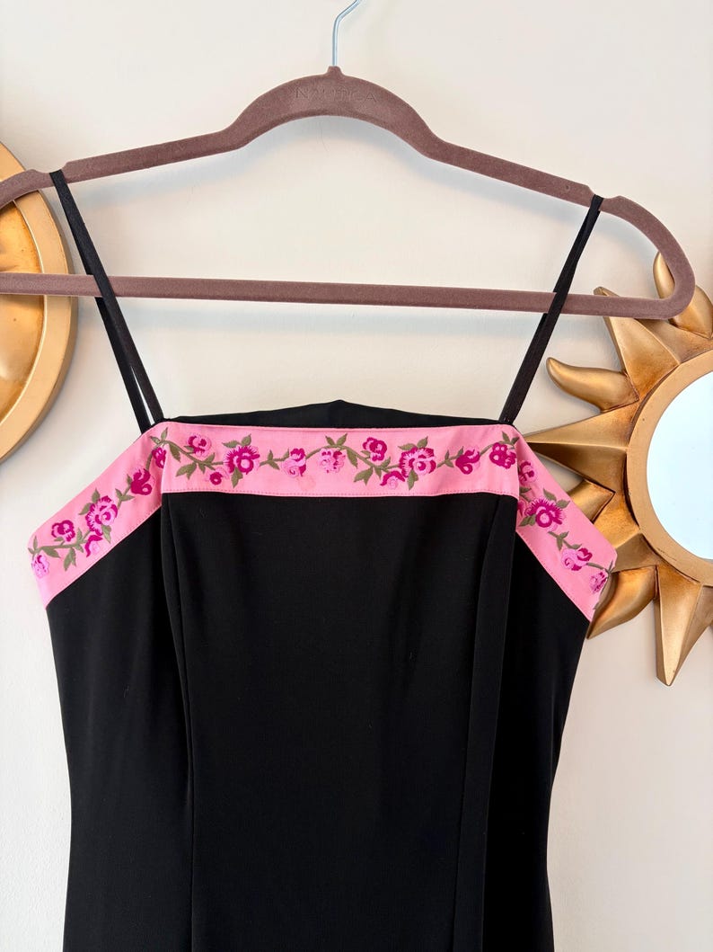 Puede incluir: Vestido negro con tirantes finos y ribete bordado floral rosa. El vestido est&aacute; colgado de una percha marr&oacute;n. El ribete presenta rosas rosas y hojas verdes.