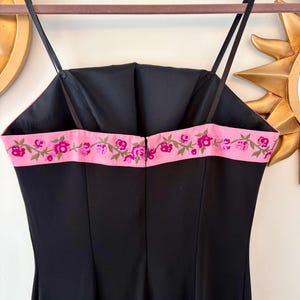 Puede incluir: Un vestido negro con tirantes finos y un ribete bordado con flores rosas. El vestido tiene un corpi&ntilde;o ajustado y un dise&ntilde;o simple y elegante. El vestido est&aacute; colgado en una percha.