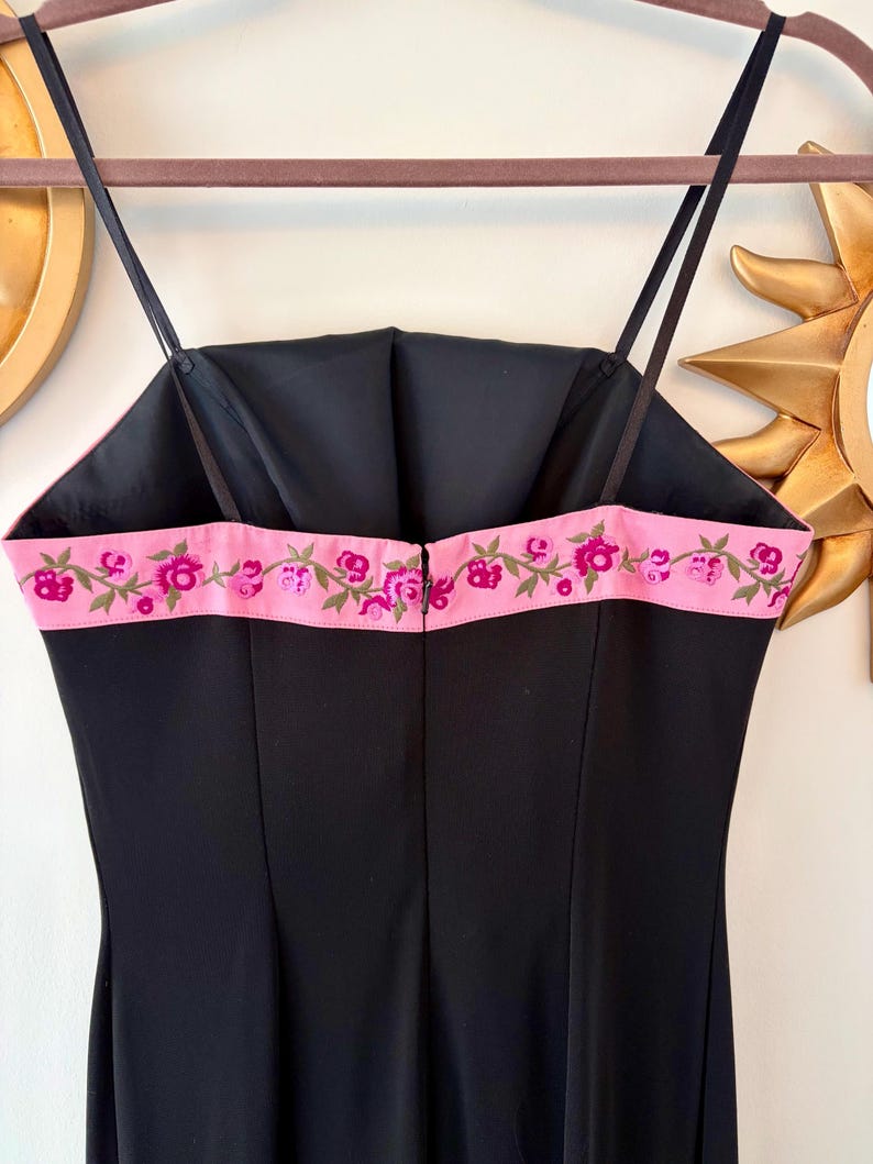 Puede incluir: Un vestido negro con tirantes finos y un ribete bordado con flores rosas. El vestido tiene un corpi&ntilde;o ajustado y un dise&ntilde;o simple y elegante. El vestido est&aacute; colgado en una percha.