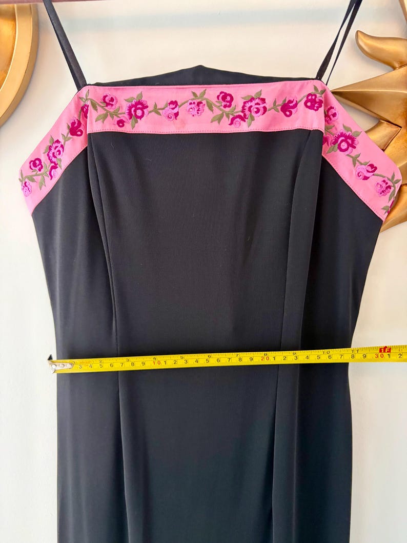 Puede incluir: Vestido negro con ribete rosa bordado con flores. El vestido tiene tirantes finos y un corpi&ntilde;o ajustado. La cintura mide aproximadamente 40 cm de ancho. El ribete presenta flores rosas y hojas verdes.