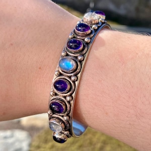 May include: Silver cuff bracelet featuring oval amethyst and moonstone gemstones. The bracelet has a detailed design with a series of bezel-set stones. The gemstones are a deep purple and iridescent blue, set against the silver metal.