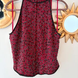 K&ouml;nnte beinhalten: Ein rotes Neckholder-Top mit hohem Ausschnitt, verziert mit schwarzen, perlenbesetzten Blumenmustern. Das Top hat eine schwarze Perlenborte und rote Tr&auml;ger. Das Kleidungsst&uuml;ck h&auml;ngt an einem Holzb&uuml;gel.