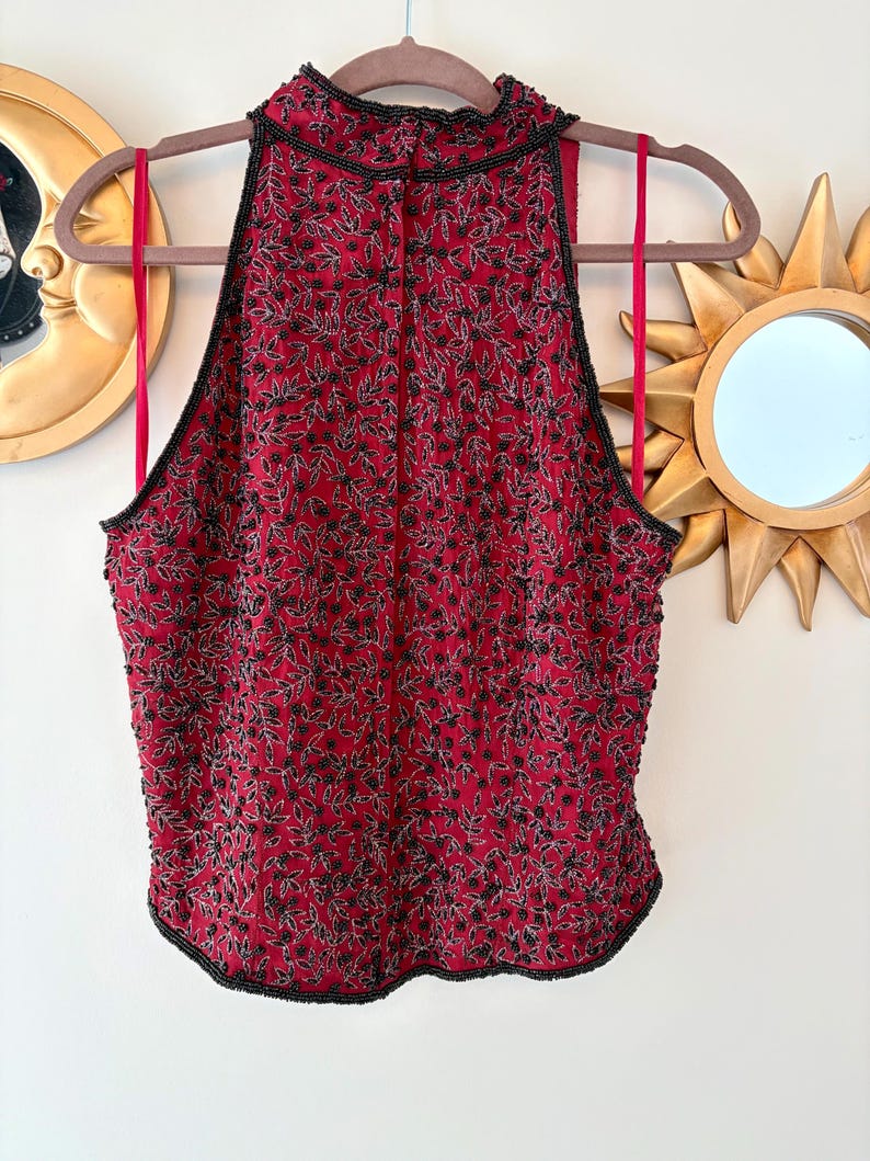 K&ouml;nnte beinhalten: Ein rotes Neckholder-Top mit hohem Ausschnitt, verziert mit schwarzen, perlenbesetzten Blumenmustern. Das Top hat eine schwarze Perlenborte und rote Tr&auml;ger. Das Kleidungsst&uuml;ck h&auml;ngt an einem Holzb&uuml;gel.