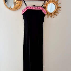 Puede incluir: Un vestido largo negro de terciopelo con tirantes finos y un ribete rosa bordado con flores. El vestido est&aacute; colgado de una percha marr&oacute;n contra una pared neutra, con espejos decorativos dorados en el fondo.