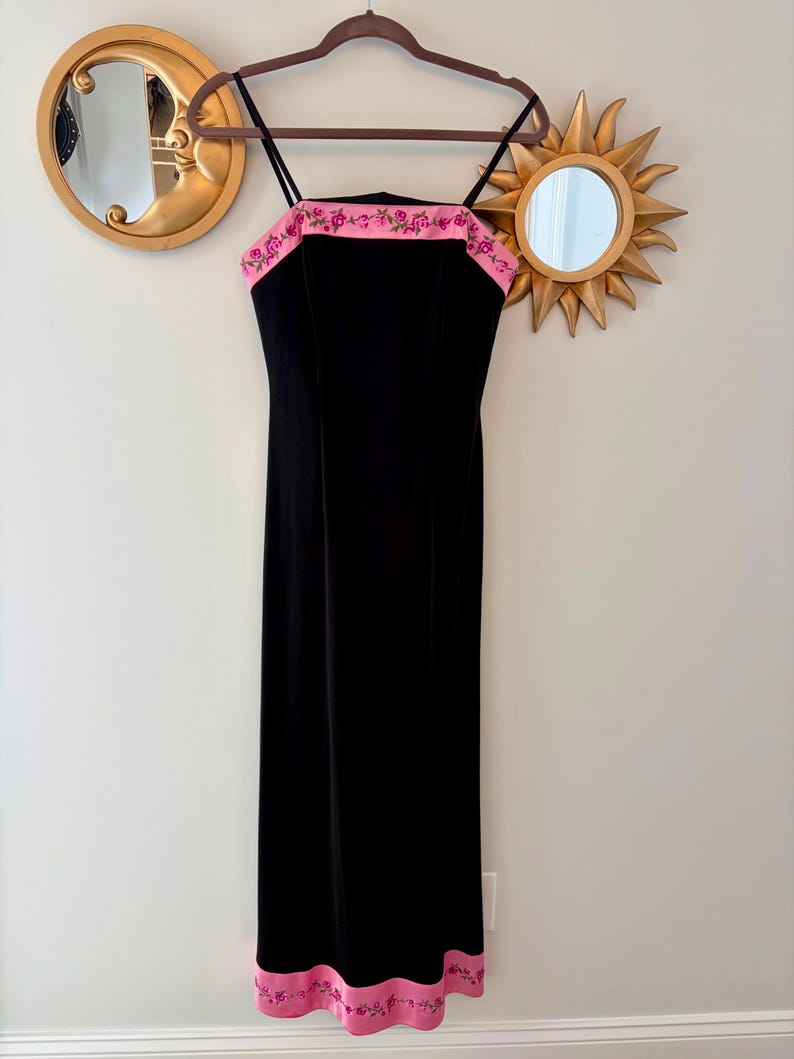 Puede incluir: Un vestido largo negro de terciopelo con tirantes finos y un ribete rosa bordado con flores. El vestido est&aacute; colgado de una percha marr&oacute;n contra una pared neutra, con espejos decorativos dorados en el fondo.