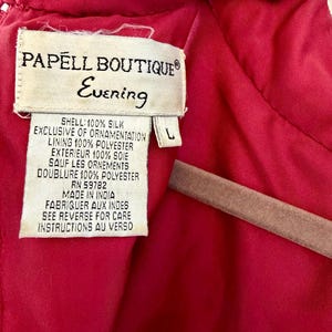 K&ouml;nnte beinhalten: Nahaufnahme eines Etiketts eines Papell Boutique Evening Kleides. Das Etikett ist cremefarben mit schwarzem Text, einschlie&szlig;lich des Markennamens und &bdquo;Evening&ldquo;. Das Kleid ist leuchtend rot, mit einem Etikett der Gr&ouml;&szlig;e L. Die Stoffzusammensetzung ist angegeben.