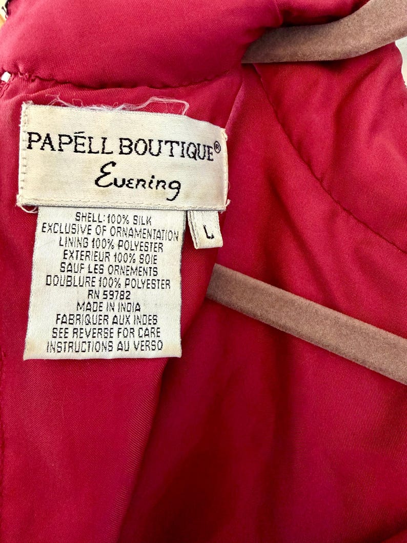 K&ouml;nnte beinhalten: Nahaufnahme eines Etiketts eines Papell Boutique Evening Kleides. Das Etikett ist cremefarben mit schwarzem Text, einschlie&szlig;lich des Markennamens und &bdquo;Evening&ldquo;. Das Kleid ist leuchtend rot, mit einem Etikett der Gr&ouml;&szlig;e L. Die Stoffzusammensetzung ist angegeben.