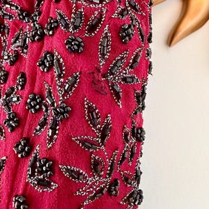 K&ouml;nnte beinhalten: Nahaufnahme eines magentafarbenen Stoffes, der mit schwarzen Perlen und silbernem Faden verziert ist. Das Design zeigt Blumen- und Blattmuster. Die Perlen sind zu Blumen geformt und umrei&szlig;en Bl&auml;tter, wodurch eine strukturierte Oberfl&auml;che entsteht.
