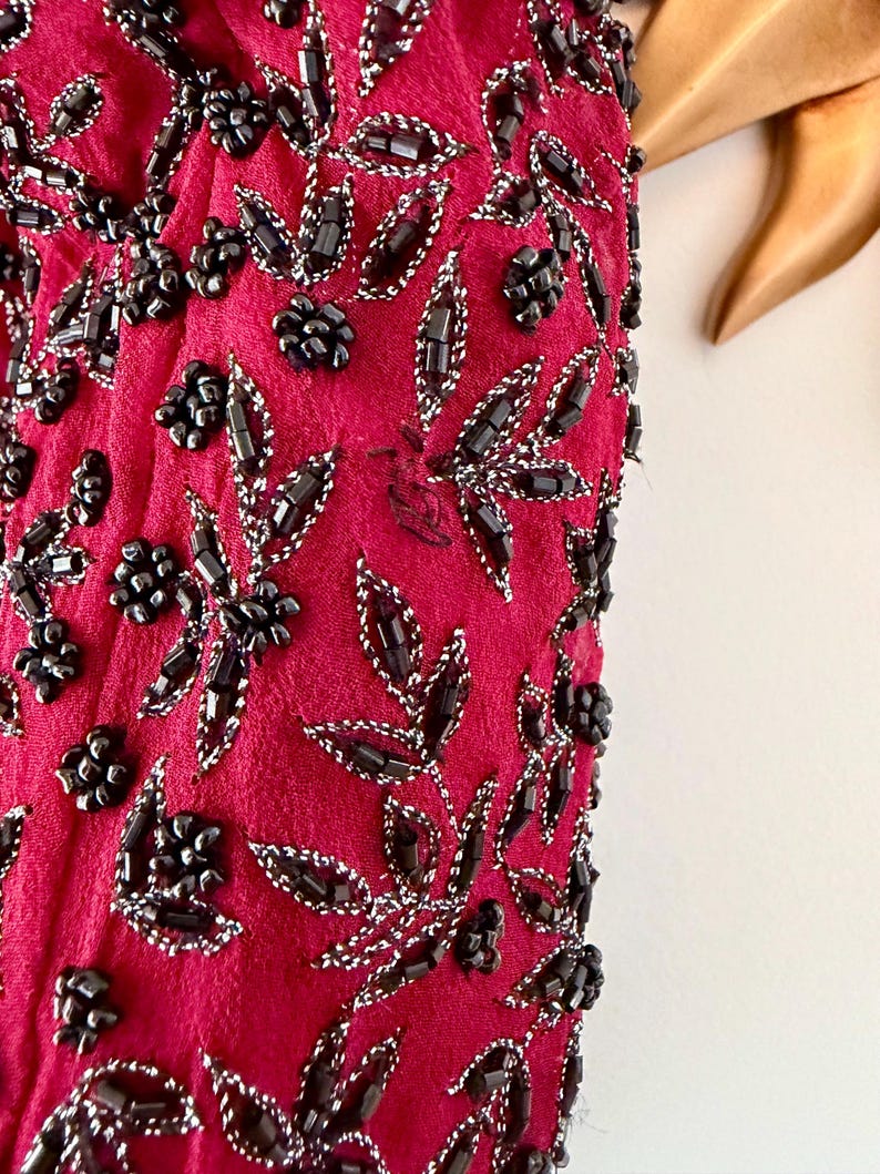 K&ouml;nnte beinhalten: Nahaufnahme eines magentafarbenen Stoffes, der mit schwarzen Perlen und silbernem Faden verziert ist. Das Design zeigt Blumen- und Blattmuster. Die Perlen sind zu Blumen geformt und umrei&szlig;en Bl&auml;tter, wodurch eine strukturierte Oberfl&auml;che entsteht.
